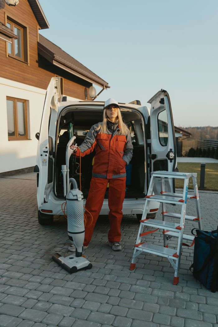 who-we-are Une agente de nettoyage en uniforme se tient à côté d’un van avec du matériel de nettoyage, devant une maison moderne au coucher du soleil.